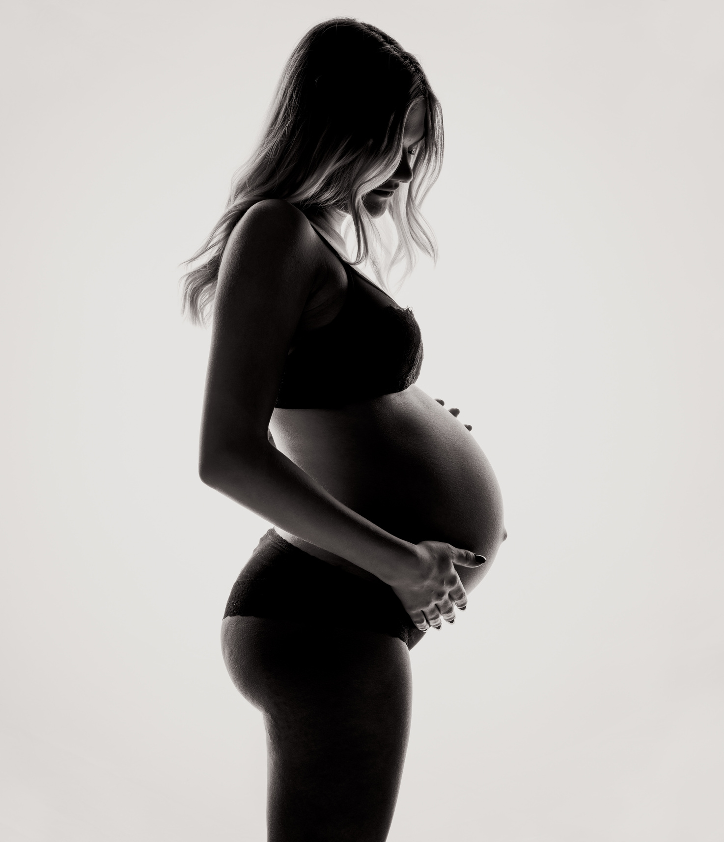 Profile of pregnant woman in underwear with hands on belly in black and white in front of white background. 

Midwife Kaleigh supports active labor in home births. 